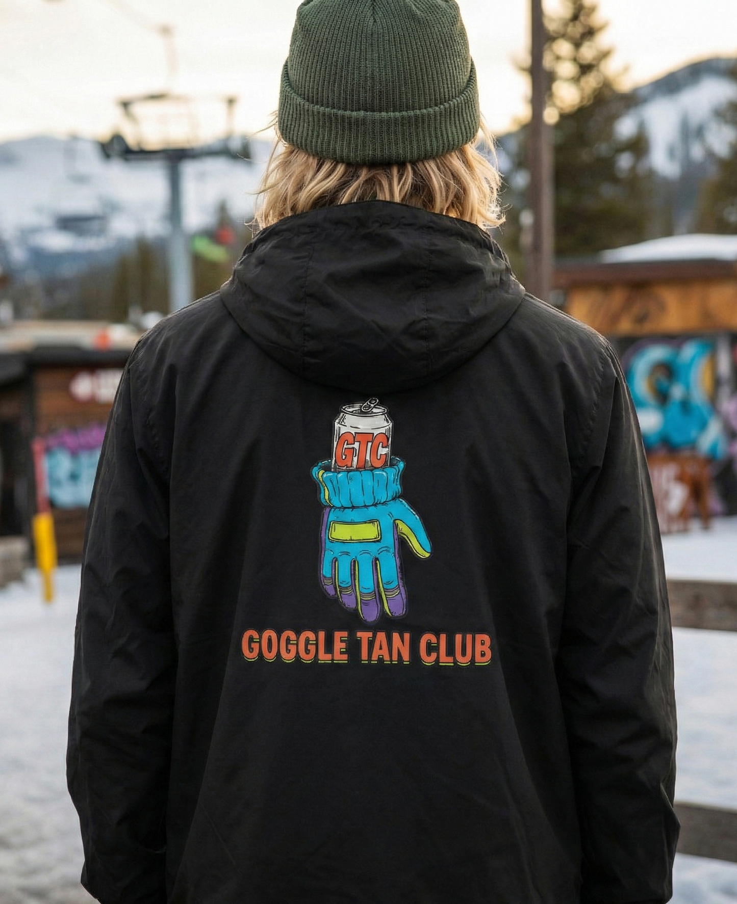 Person wearing a black jacket with 'Goggle Tan Club' design in a snowy outdoor setting