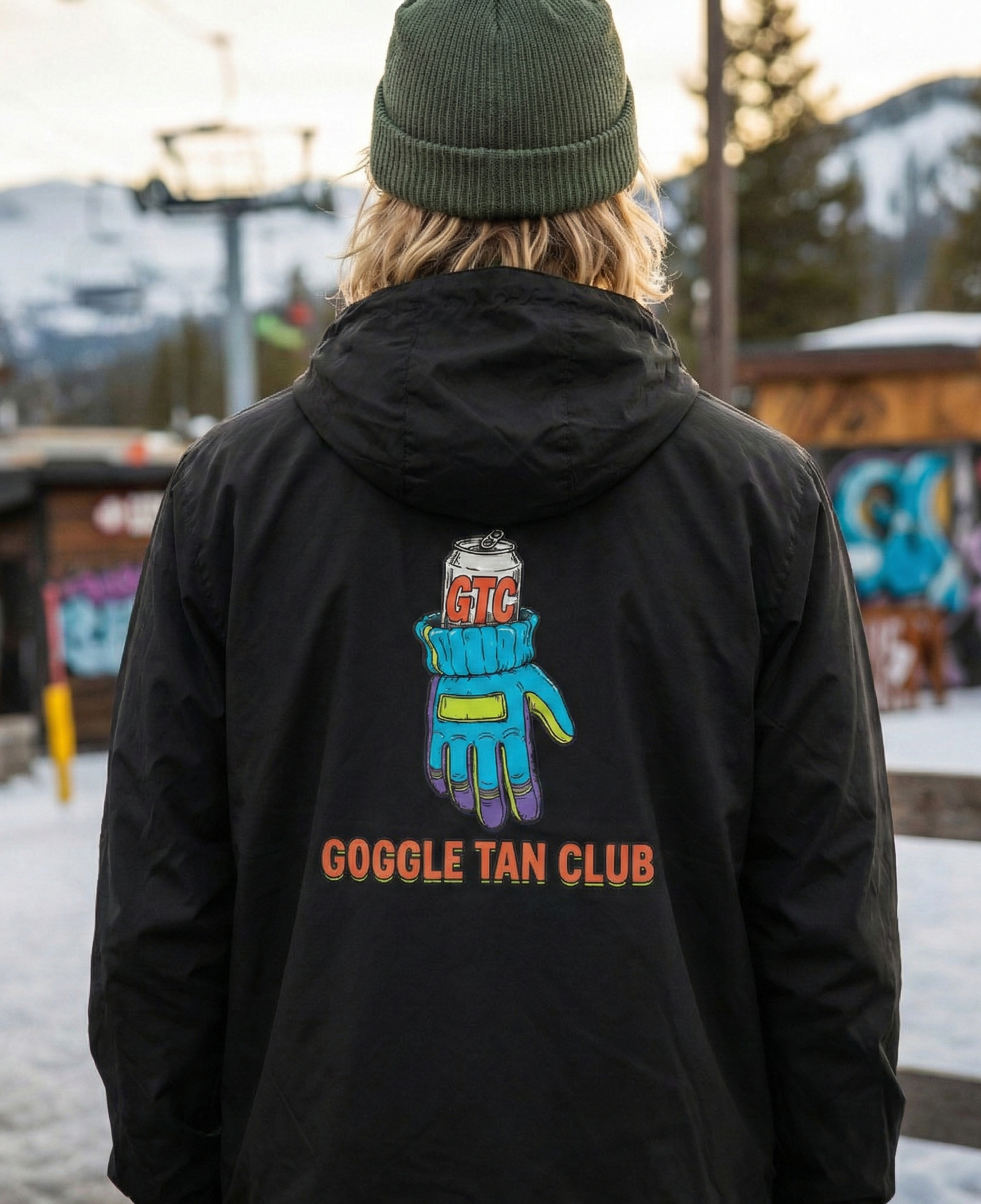 Person wearing a black jacket with 'Goggle Tan Club' design in a snowy outdoor setting