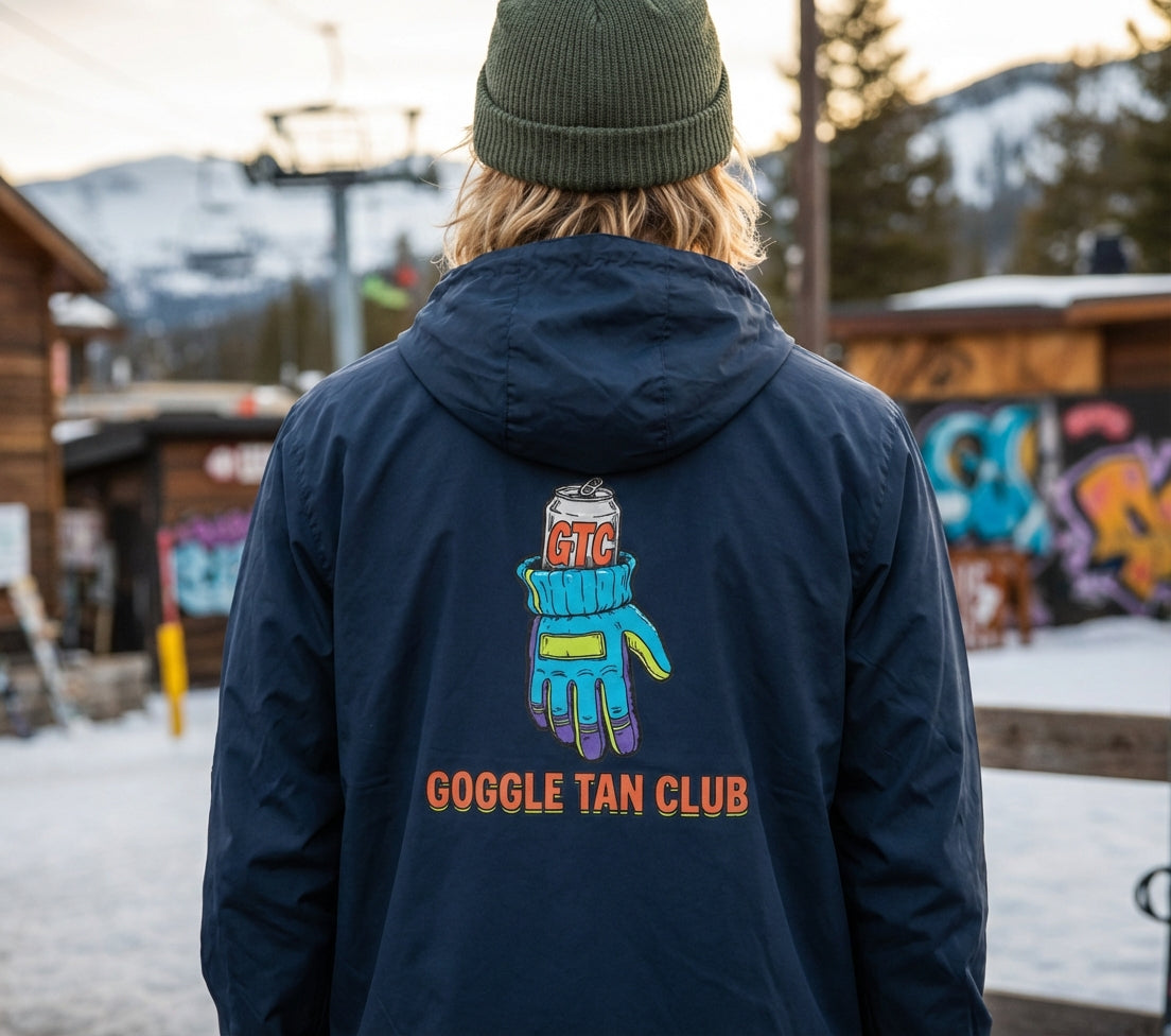 Person wearing a navy blue jacket with 'Goggle Tan Club' logo in a snowy outdoor setting